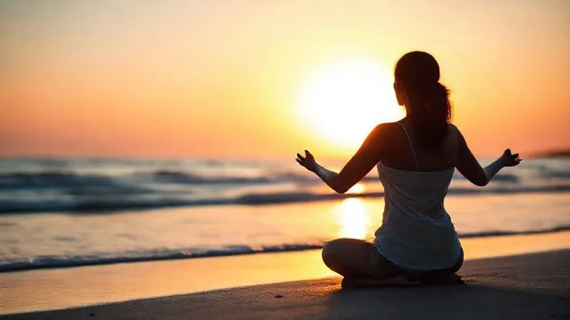 A photorealistic, professional photography shot of a person (gender-neutral, 30s) meditating peacefully on a tranquil beach at sunrise, silhouetted against the vibrant sky. The sharp focus is on the person's calm posture, with gentle waves lapping the shore and a soft, warm glow in the background. Cinematic lighting, depth of field, 8K hyper-detailed, shot on a high-end DSLR, conveying deep introspection and a sense of inner peace.