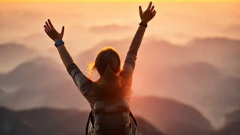 A photorealistic, professional photography shot of a solo female traveler (30s, diverse ethnicity) standing victoriously at a mountain summit, arms raised in celebration, gazing at a panoramic view. The sun is setting, casting a warm glow. Sharp focus on her joyful expression and silhouette, with the vast, blurred mountain range in the background. Cinematic lighting, depth of field, 8K hyper-detailed, shot on a high-end DSLR, conveying a powerful sense of accomplishment and self-worth.