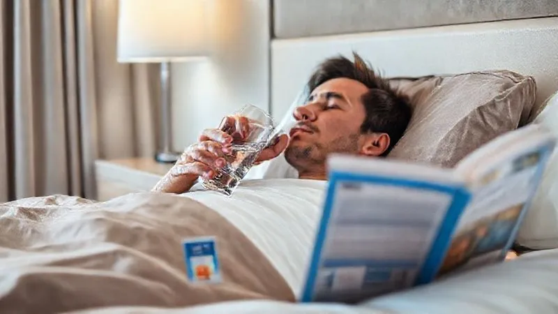 A photorealistic, professional photography, 8K, cinematic lighting, sharp focus, depth of field, shot on a high-end DSLR, showing a traveler resting comfortably in a clean hotel room, sipping from a glass of water with an ORS packet nearby, a travel guide open but unread, conveying a sense of calm recovery after a minor setback.