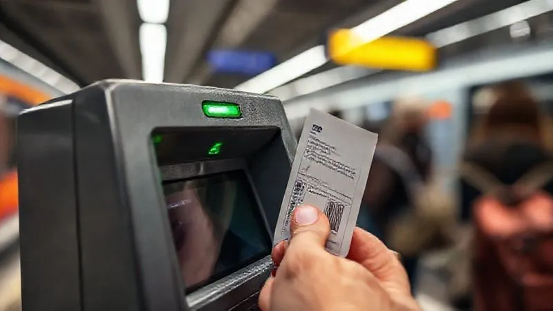 photorealistic, professional photography, 8K, cinematic lighting, sharp focus, depth of field, shot on a high-end DSLR. A close-up shot of a hand inserting a paper public transport ticket into a small validation machine at a train station entrance. The machine has a green light indicating success, with blurred commuters in the background, emphasizing a crucial, often overlooked step in city travel.