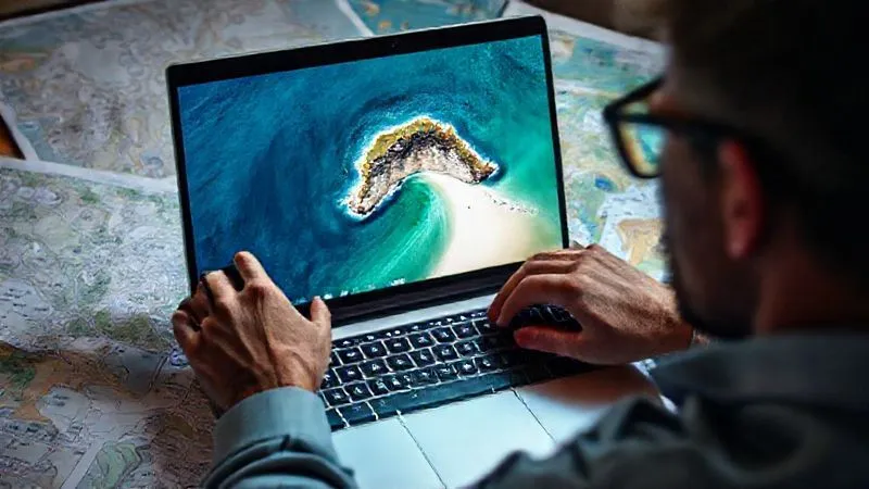 A person intently focused on a laptop screen, surrounded by maps and travel guides, deep diving into online forums and satellite imagery to uncover a hidden beach. The screen shows a detailed satellite view of a coastline with a tiny, isolated cove. Photorealistic, professional photography, 8K, cinematic lighting, sharp focus, depth of field, shot on a high-end DSLR.