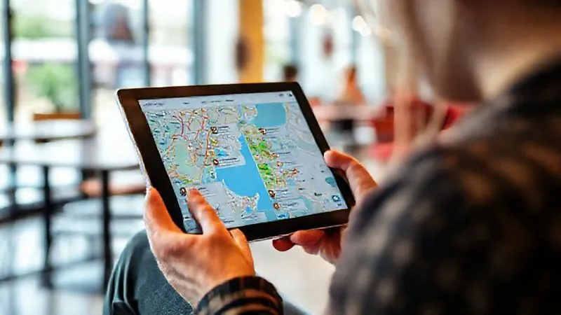 A person meticulously researching travel destinations on a tablet, with accessibility icons highlighted on a map, sitting in a bright, modern cafe. The tablet screen shows detailed maps and user reviews for accessible points of interest. Photorealistic, professional photography, 8K, cinematic lighting, sharp focus, depth of field.