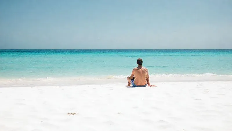 A photorealistic shot of a lone figure sitting peacefully on a genuinely secluded, dazzling white sand beach, gazing out at the calm, crystal-clear turquoise ocean. The beach is pristine, with no other people or signs of development. The atmosphere is one of profound serenity and solitude. 8K, professional photography, cinematic lighting, sharp focus on the figure and the foreground, depth of field blurring the distant horizon.