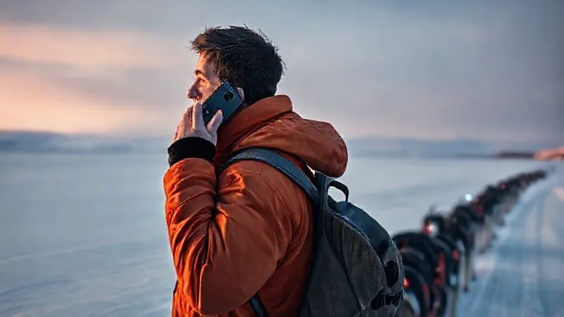 Photorealistic, professional photography of a musher standing next to their sled, holding a satellite phone to their ear, looking out at a vast, snow-covered landscape. A faint signal light from a SPOT device is visible on their pack. Cinematic lighting, sharp focus on the musher's determined expression, depth of field showing the scale of the wilderness. 8K hyper-detailed, shot on a high-end DSLR, conveying a sense of urgency and preparedness.