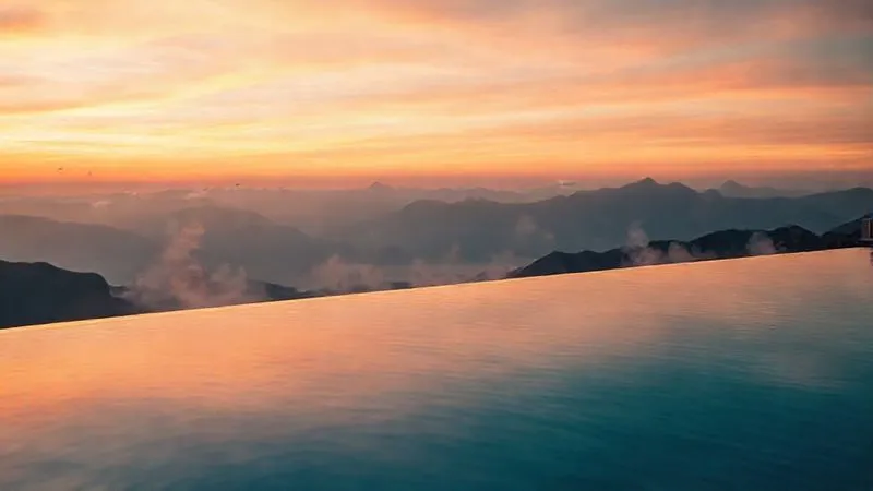 Photorealistic professional photography of a luxury wellness resort's infinity pool overlooking a serene mountain range at sunrise. The water is perfectly still, reflecting the vibrant sky, with steam gently rising. Sharp focus on the tranquil scene, depth of field blurring distant peaks. 8K hyper-detailed, cinematic lighting.